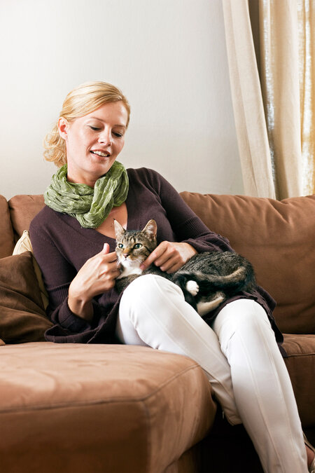 Auf einem braunen Sofa sitzt eine blonde Frau, auf deren Schloss eine Katze liegt und sich von ihr streicheln lässt.