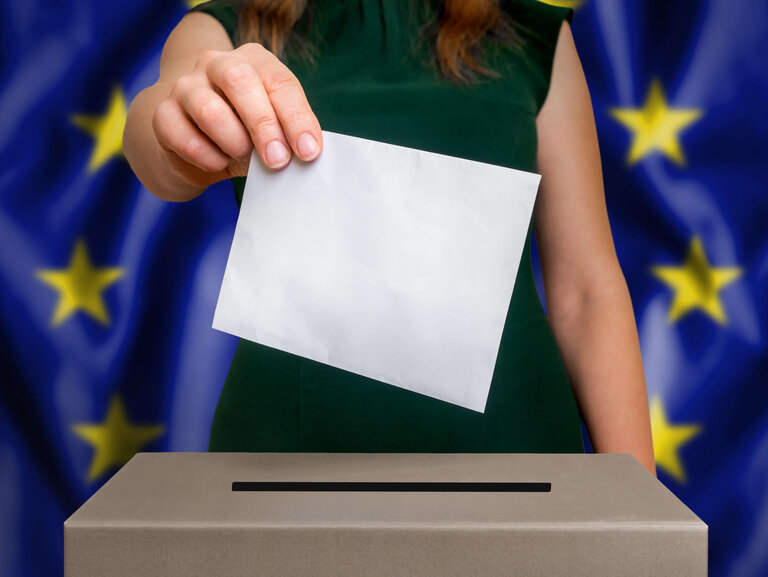 Hand wirft Wahlumschlag in Wahlurne vor Europaflagge